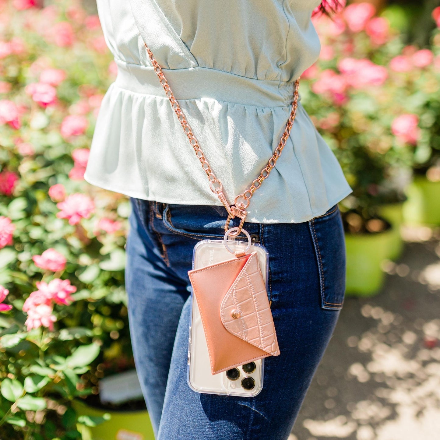 Stylish leather keychain wallet in mini envelope design. Shown connected to a crossbody strap in rose gold. The crossbody bag strap is holding a phone.