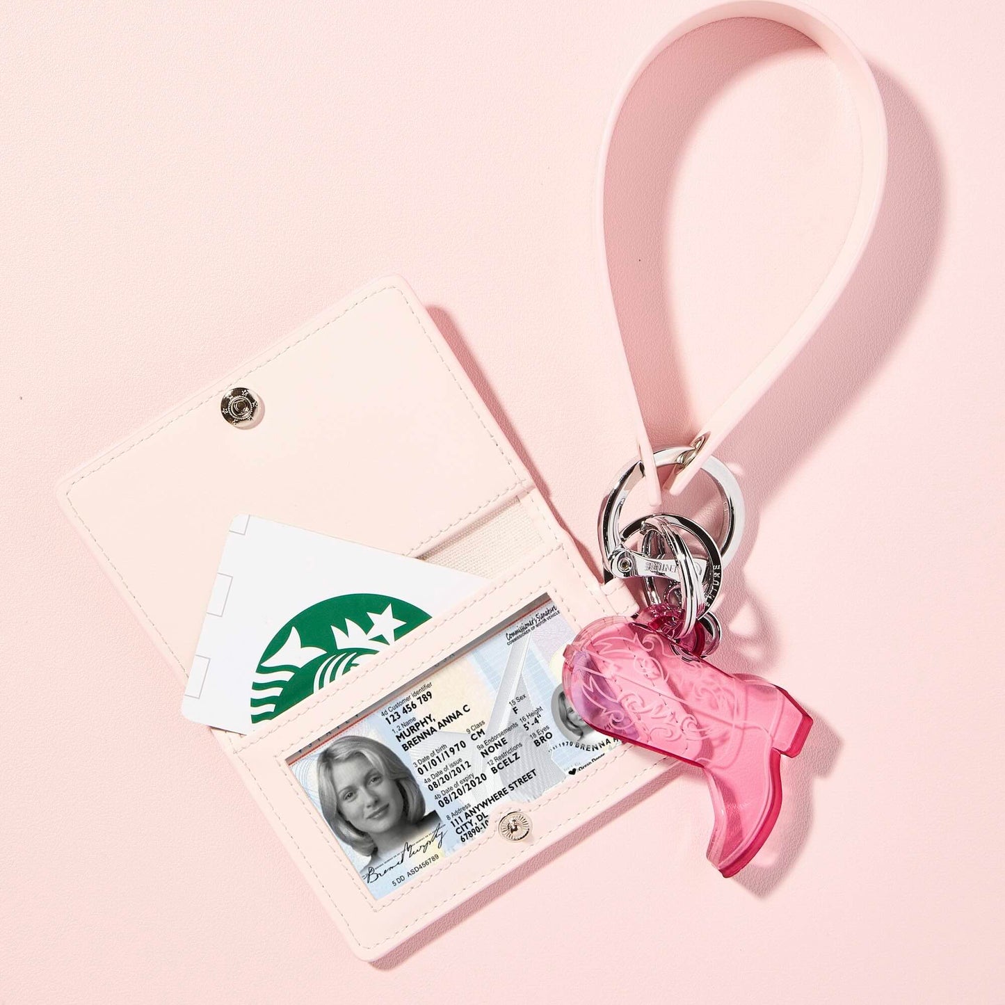 Light pink ID case with a clear ID window and Starbucks card, attached to a pink leather wristlet strap with a pink acrylic cowboy boot keychain and silver hardware, styled on a pastel pink background.