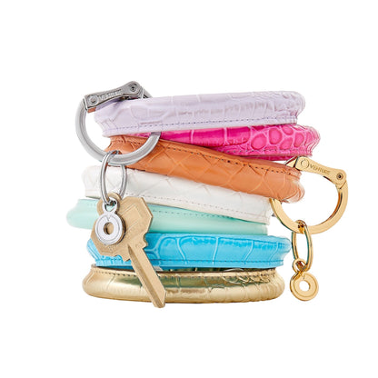A stack of Big O Key Rings in braided leather and croc embossed leather. Lavender croc, pink topaz croc, in the saddle braided, white leather braided, mint leather, peacock croc embossed leather, solid gold croc embossed leather.