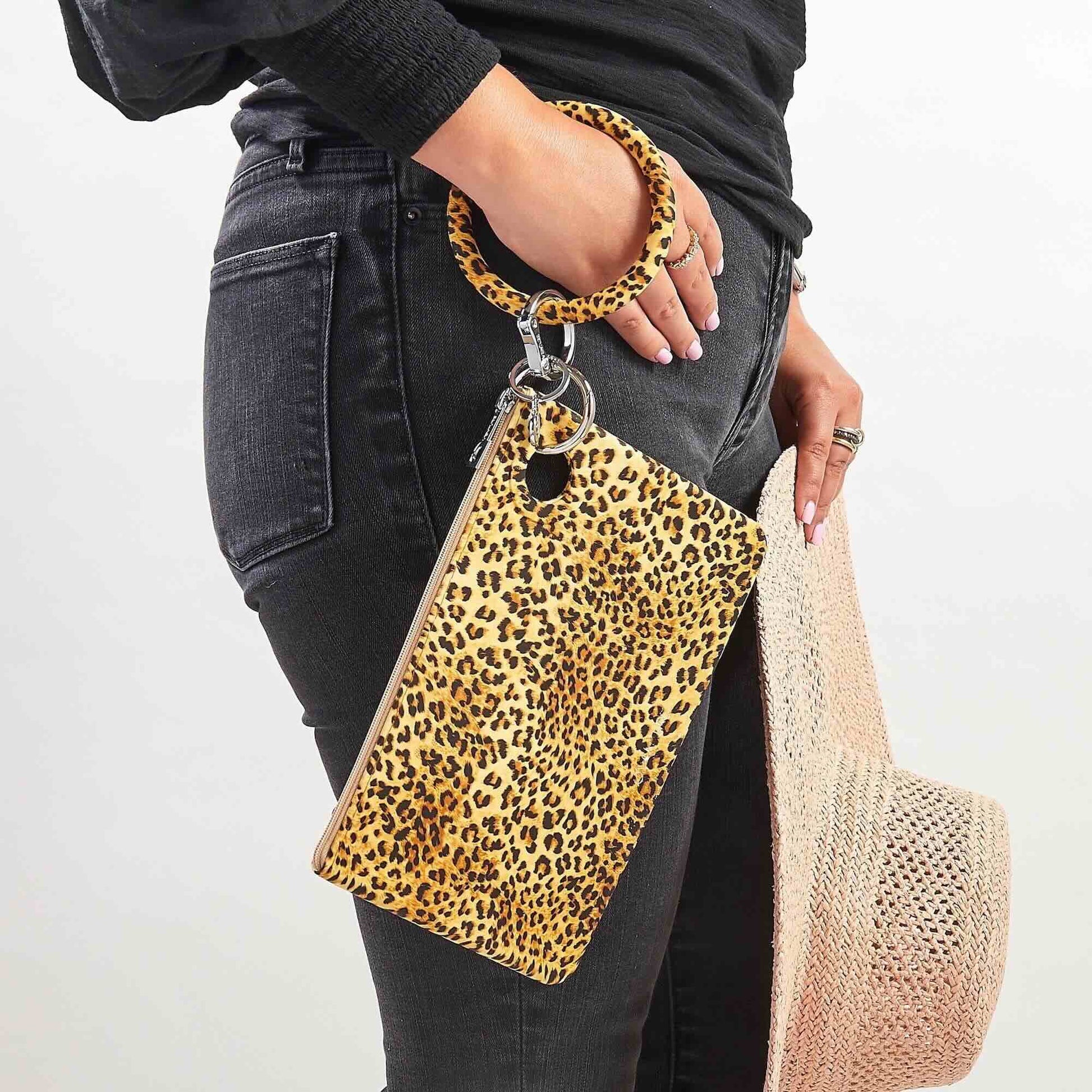 Woman wearing black jeans and a black top, holding a leopard-print silicone Big O® Key Ring with a matching large pouch wristlet and a woven straw hat, styled for a casual hands-free look.