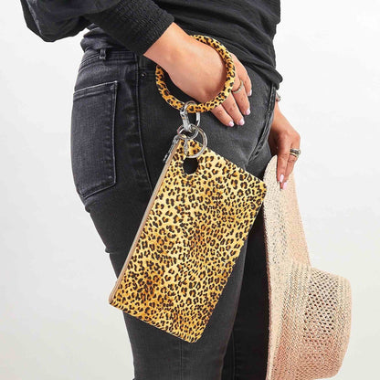 Woman wearing black jeans and a black top, holding a leopard-print silicone Big O® Key Ring with a matching large pouch wristlet and a woven straw hat, styled for a casual hands-free look.
