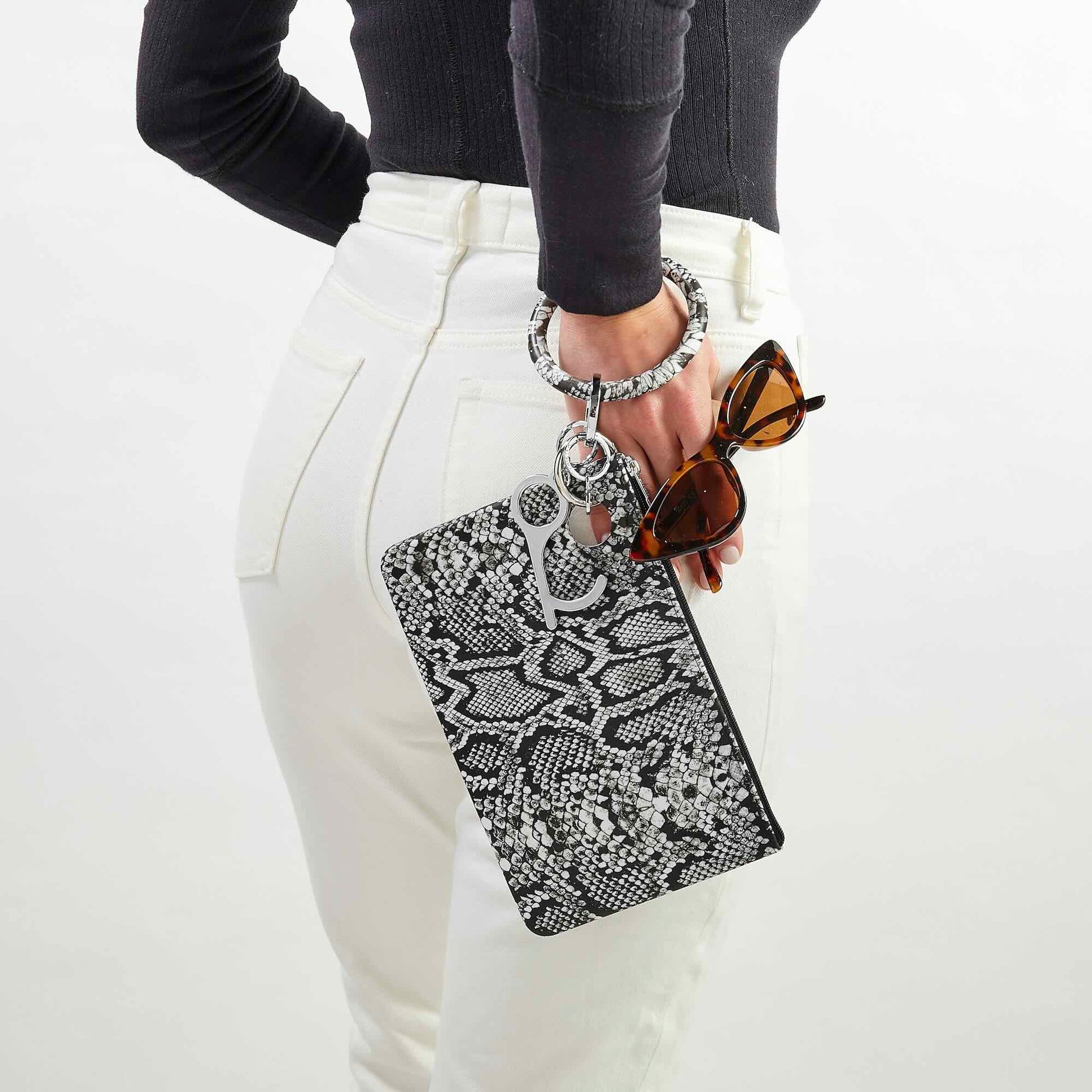 Woman wearing white jeans and a black top, holding a black and white snakeskin-print silicone Big O® Key Ring with a matching silicone pouch wristlet, sunglasses, and key essentials for a chic hands-free look.